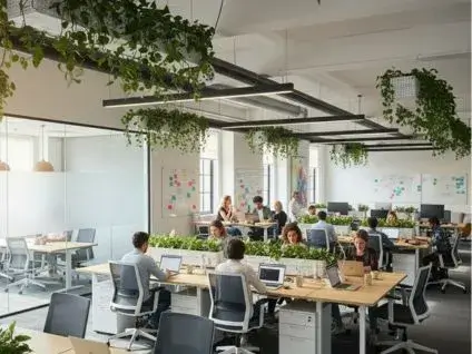 Nowoczesne biuro typu open space, w którym pracownicy różnych płci i narodowości siedzą przy długich stołach, używając laptopów i współpracując. W przestrzeni widoczne są rośliny, tablice z notatkami i duże okna, tworzące jasne i dynamiczne środowisko pracy.