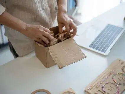 Na załączonym zdjęciu widać dłonie osoby pakującej małe, tekturowe pudełko. W środku pudełka znajduje się materiał wypełniający, a na biurku obok leży laptop i akcesoria.