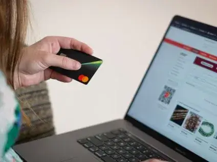 Młoda kobieta w jasnej bluzce trzyma kartę płatniczą MasterCard w dłoni, przygotowując się do wpisania danych na otwartym laptopie. Na ekranie widoczna jest strona sklepu internetowego.