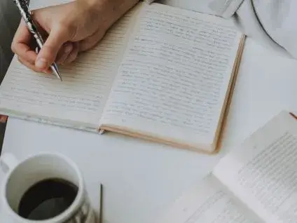 Osoba pisząca w otwartym notatniku, obok kubek z kawą.